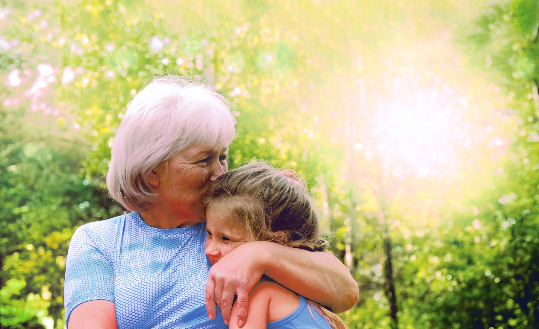 An older woman gently hugs a child in a sunlit park surrounded by trees and soft greenery
